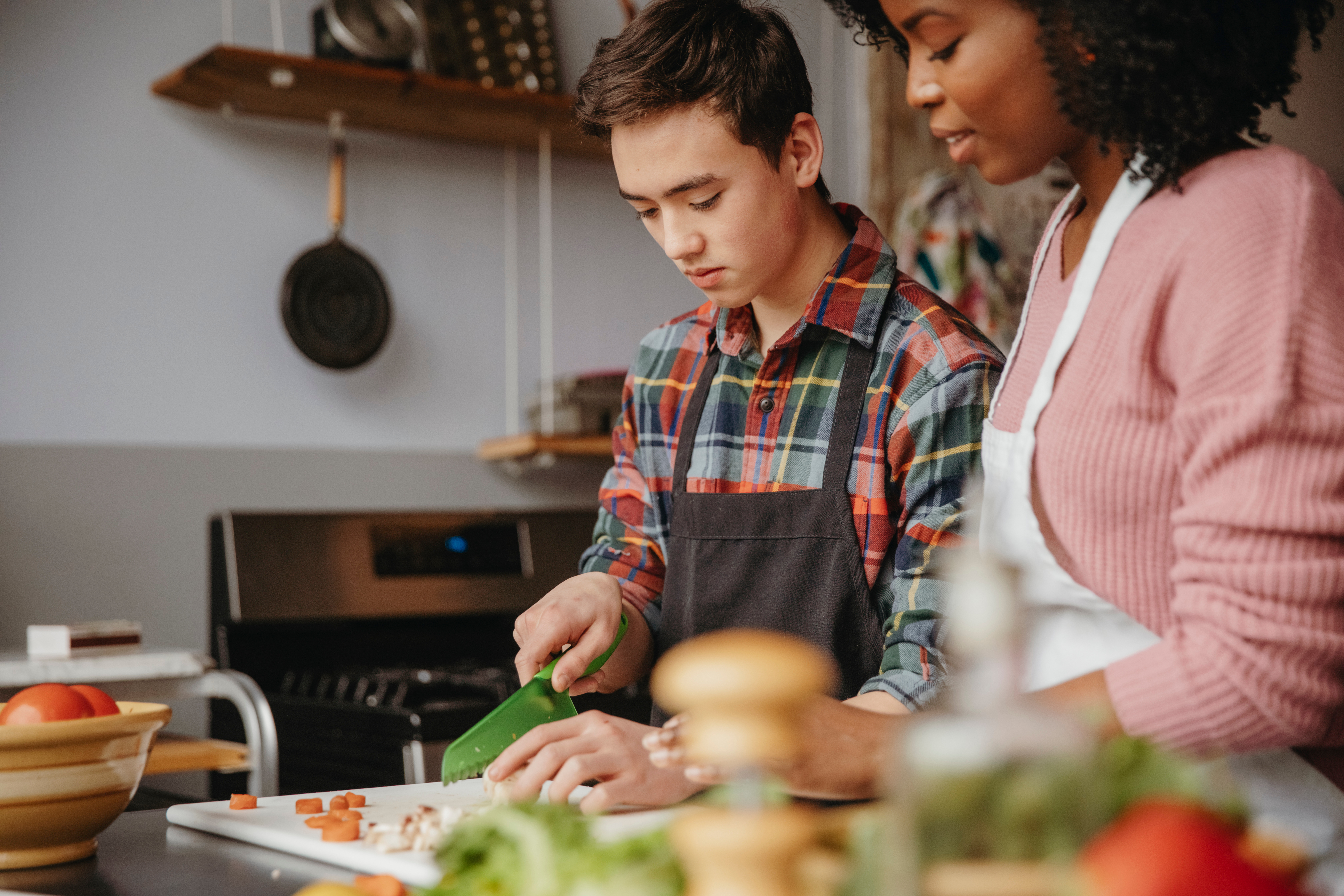 Image depicts a woman helping a young person with basic knife skills in the kitchen.