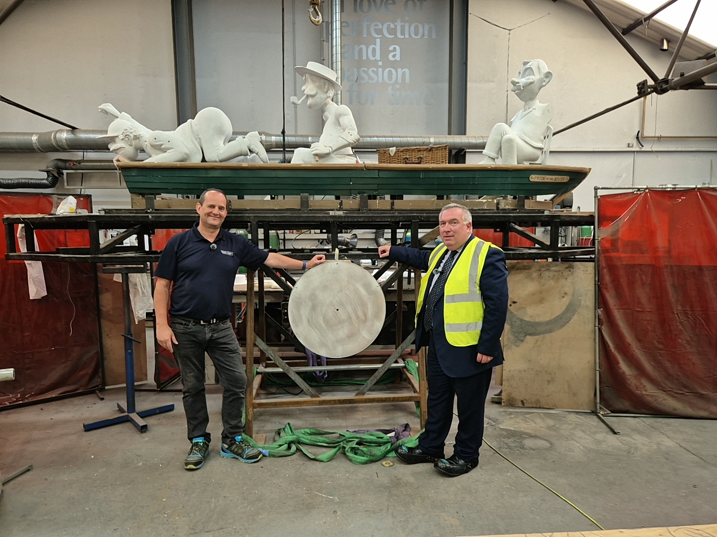Cllr Adrian Andrew standing in front of the three men in a boat in the workshop with Dean Allen