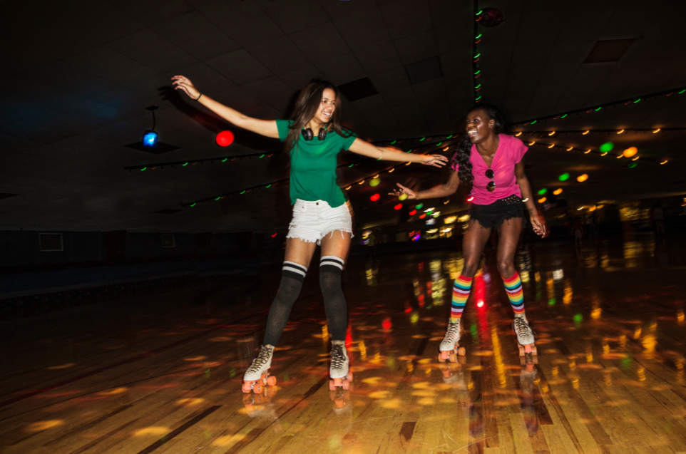 2 girls with bright tops and short with roller skates on in a darkened sporthall with disco lights skating
