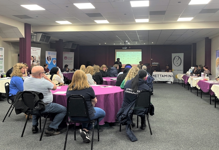 Image shows a conference room with various information stalls, round tables and attendees.