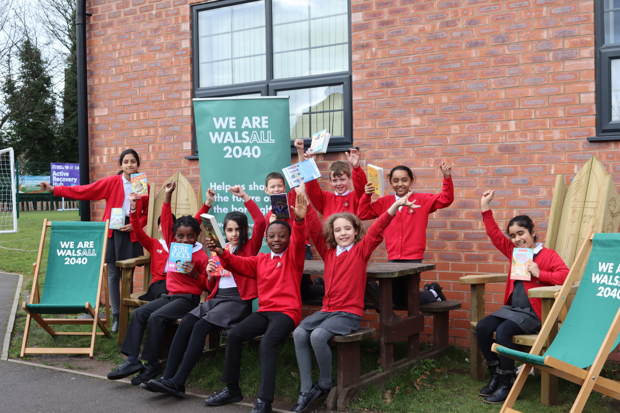 A group of school children in red jumpers sitting outside holding books and cheering