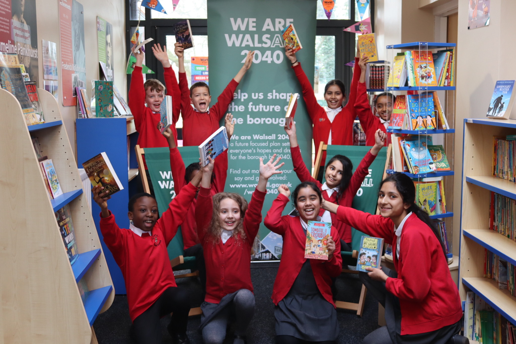 A group of school children in red jumpers sitting in a library holding books and cheering