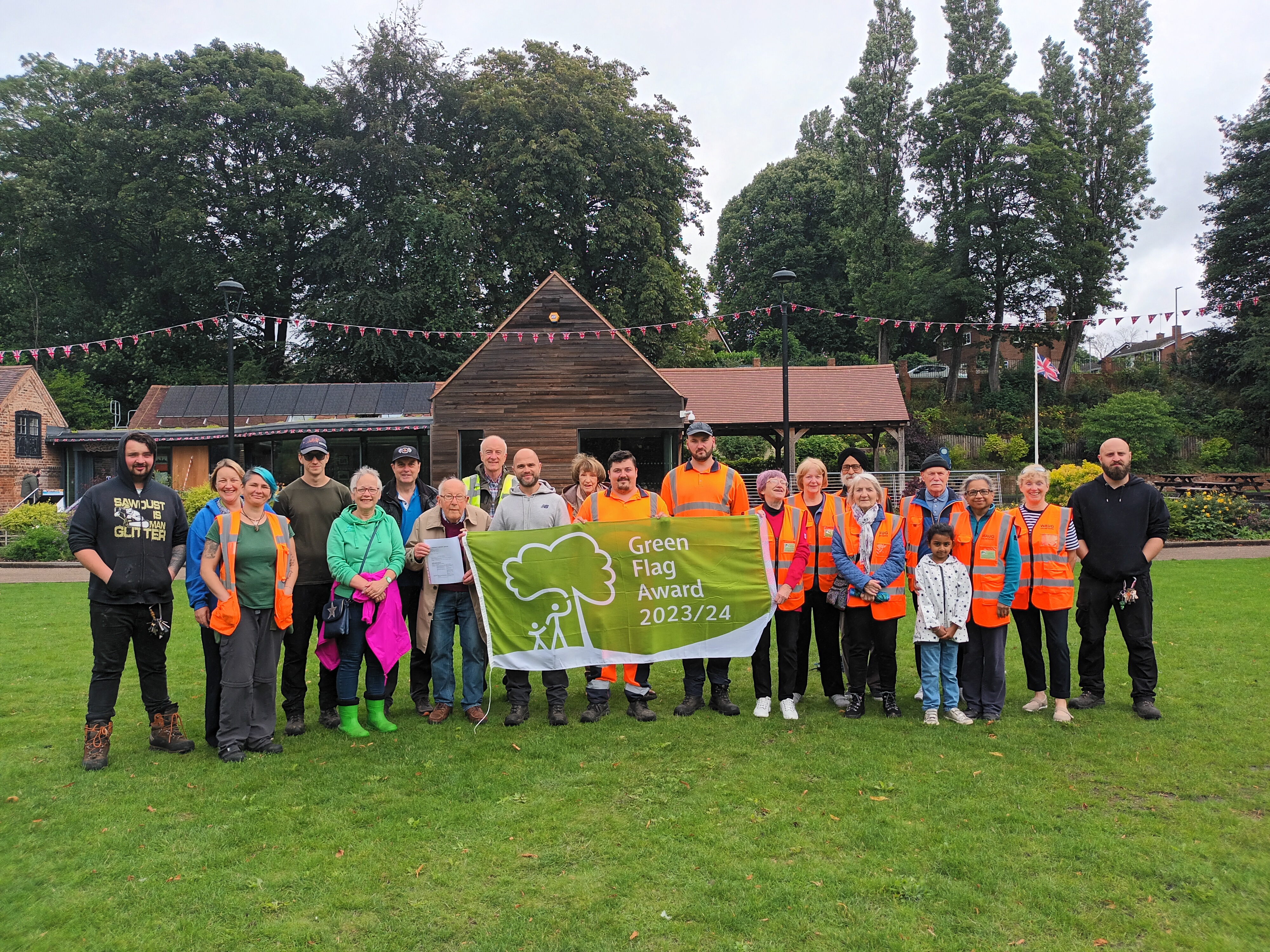 Image depicts a large group of people holding the Green Flag Award 2023/24 flag outside Walsall Arboretum Visitor Centre.