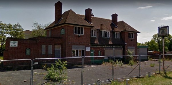 A derelict red brick pub called the Brown Jug sits vacant behind a metal fence. The lower windows are boarded up and the tarmac at the front is dotted with overgrown weeds