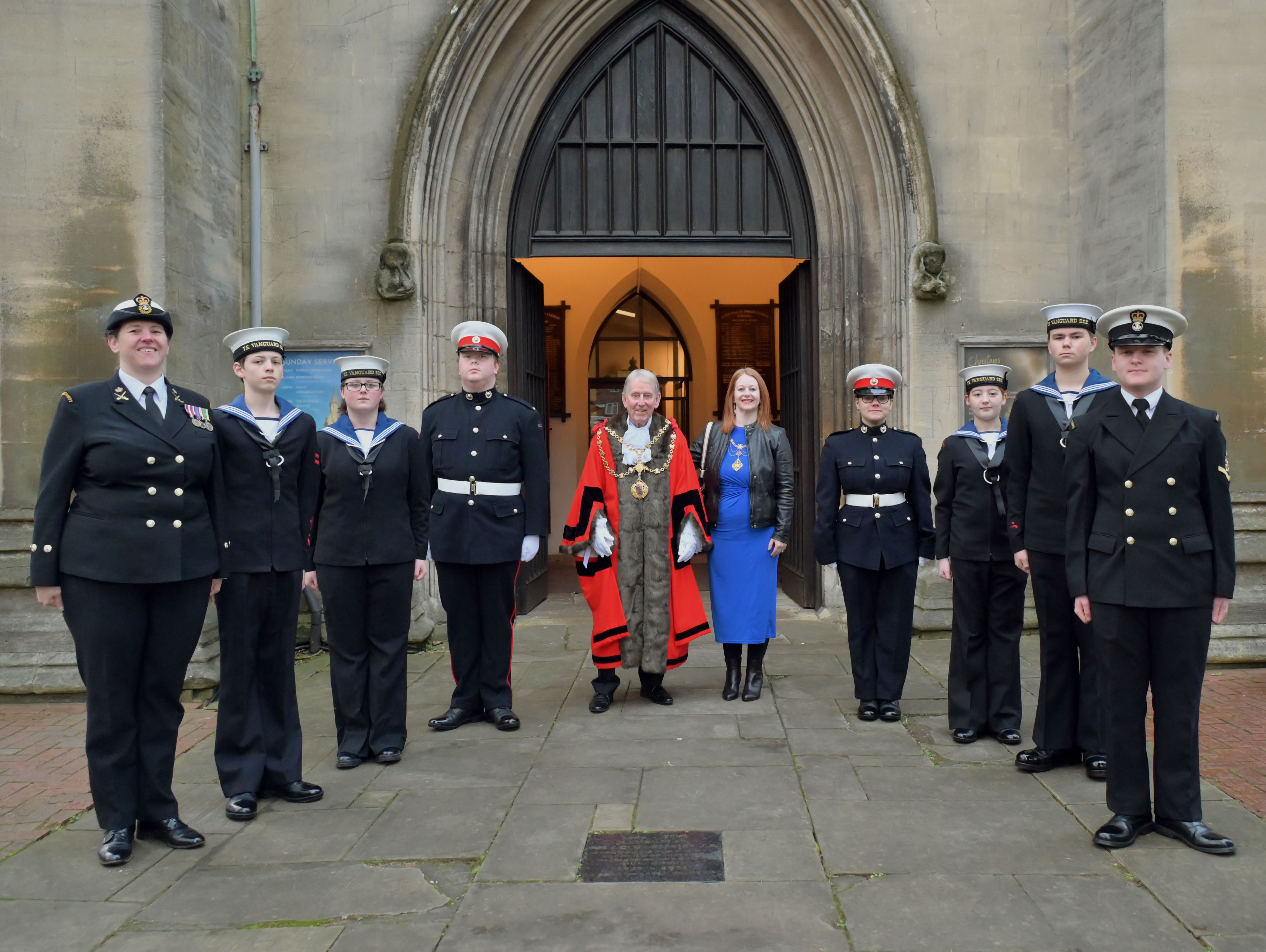 The mayor of walsall in red mayoral robes, next to the mayoress in a royal blue dress, stand smiling in the middle of a group of marine cadets in black uniforms outside a church entrance.