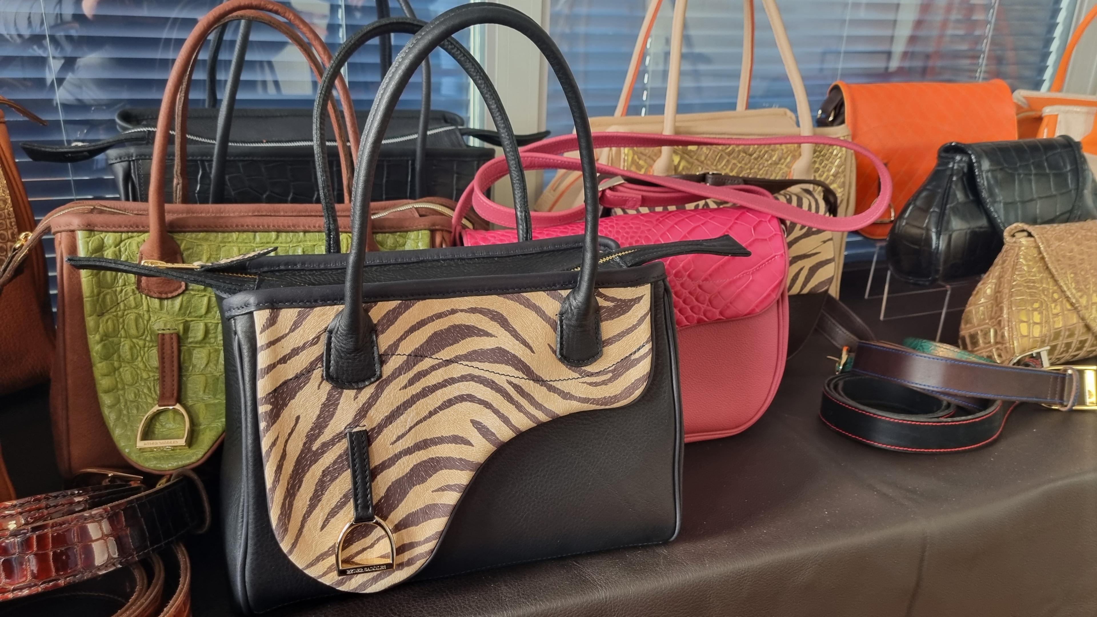 A table full of leather handbags in various colours. In front is a black bag with a zebra print top, behind it is a bright pink bag