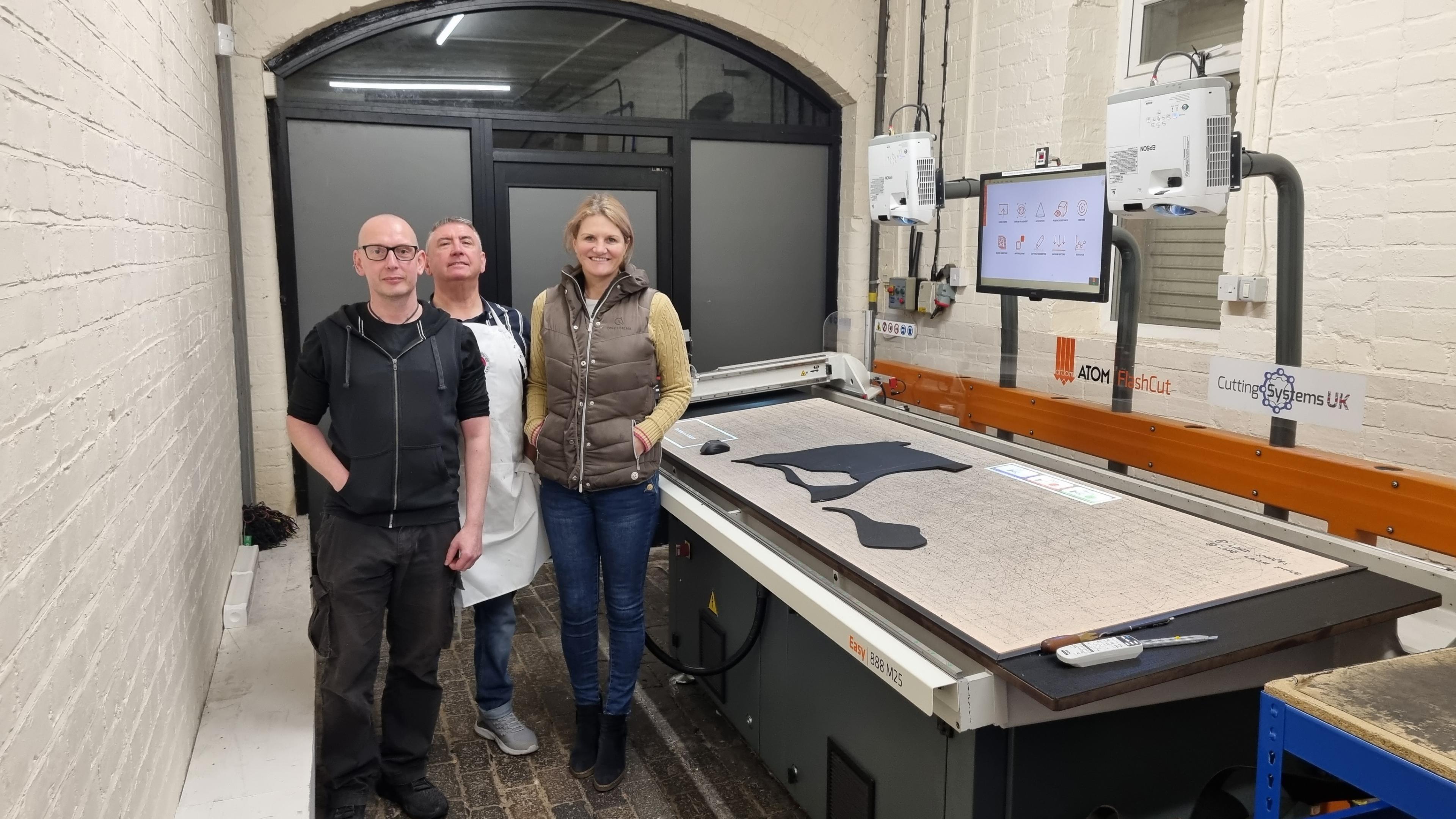 Three people standing beside a large CNC cutting machine, on which is a piece of black leather that has just been cut into shape by the machine