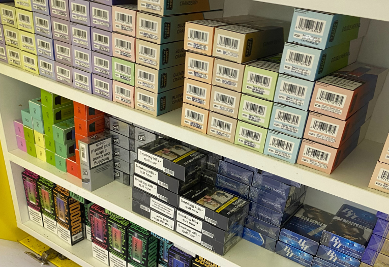 Image depicts illicit cigarettes and vapes concealed underneath a counter across three shelves.