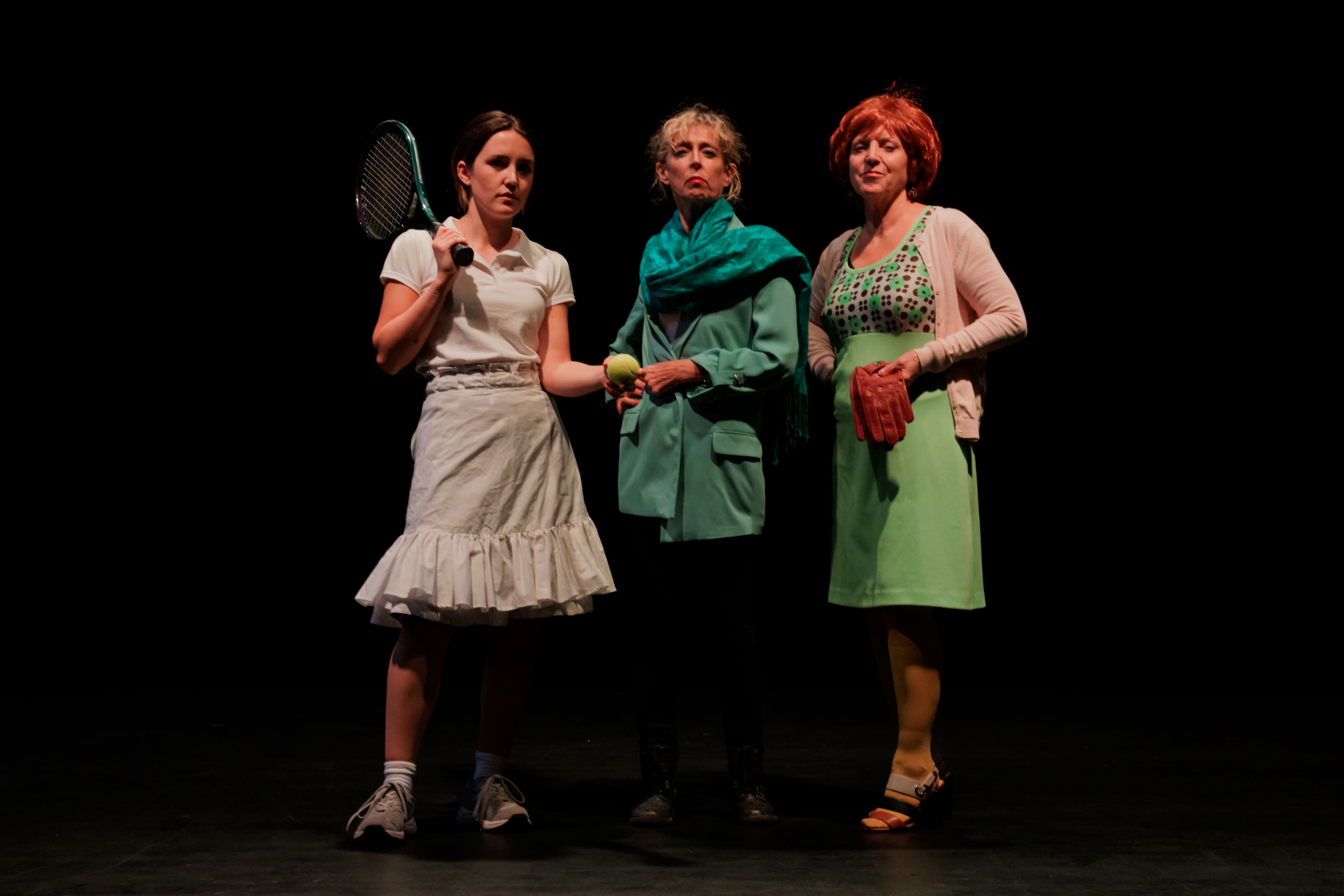 Three women, one wears a tennis outfit, one a green jacket and one a 1960s style dress