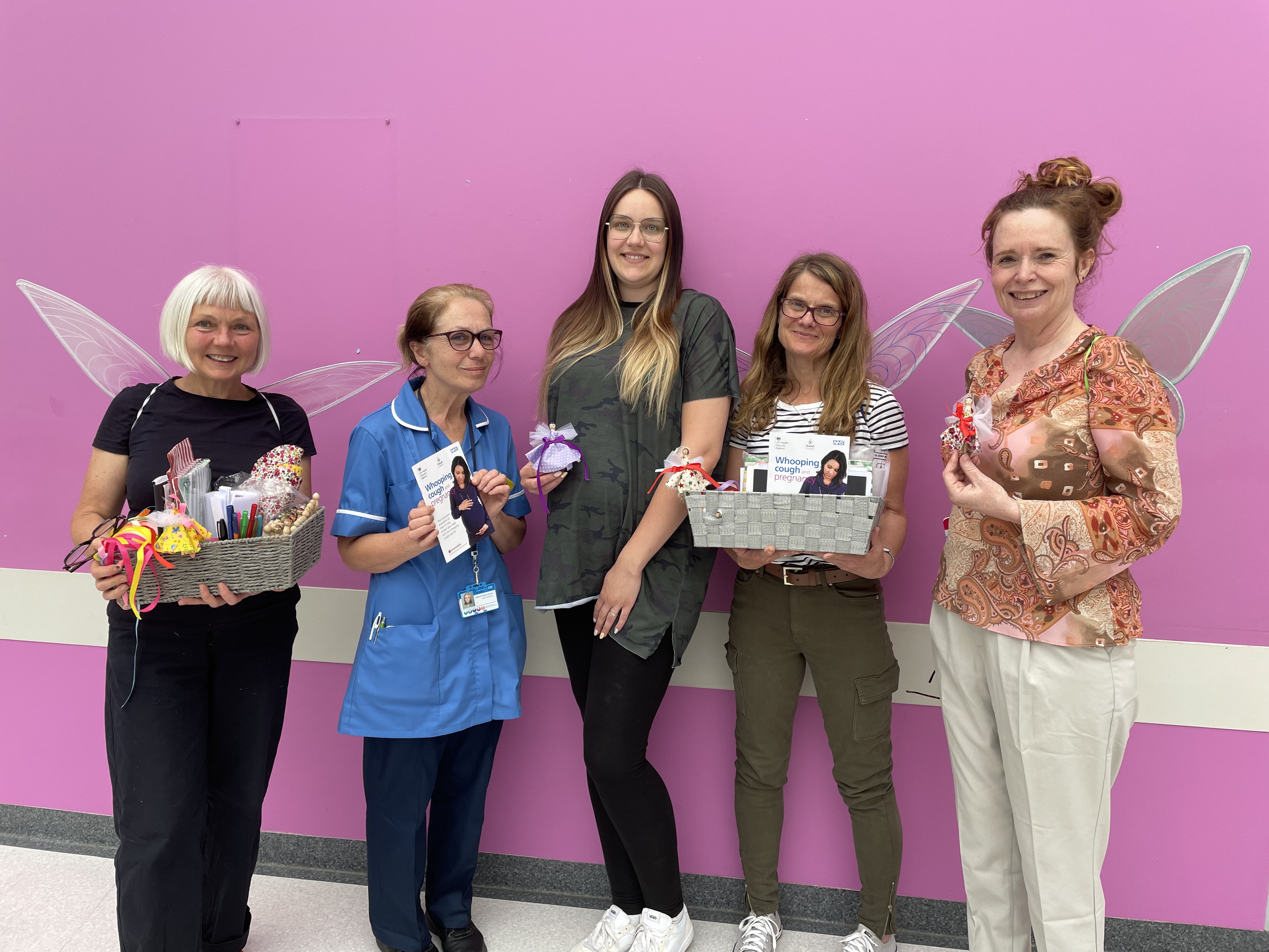 Image depicts five women wearing fairy wings, holding a basket, whooping cough booklets and a handmade fairy.
