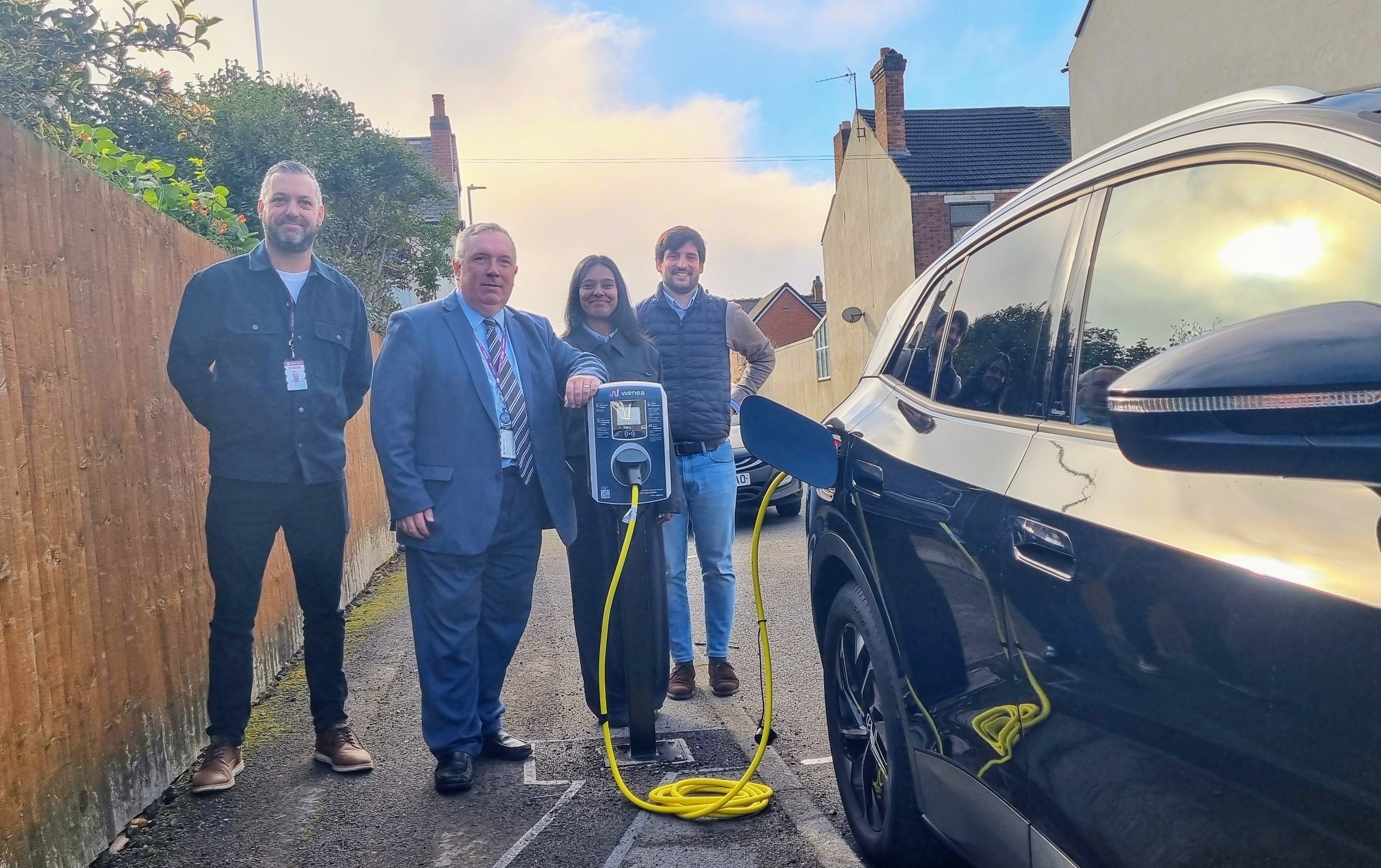 Four people stand next to a black car plugged into an electric vehicle chargepoint 