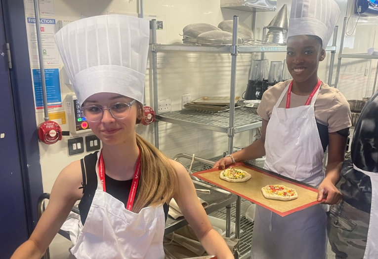 Image depicts two young people holding baking trays of pizza.