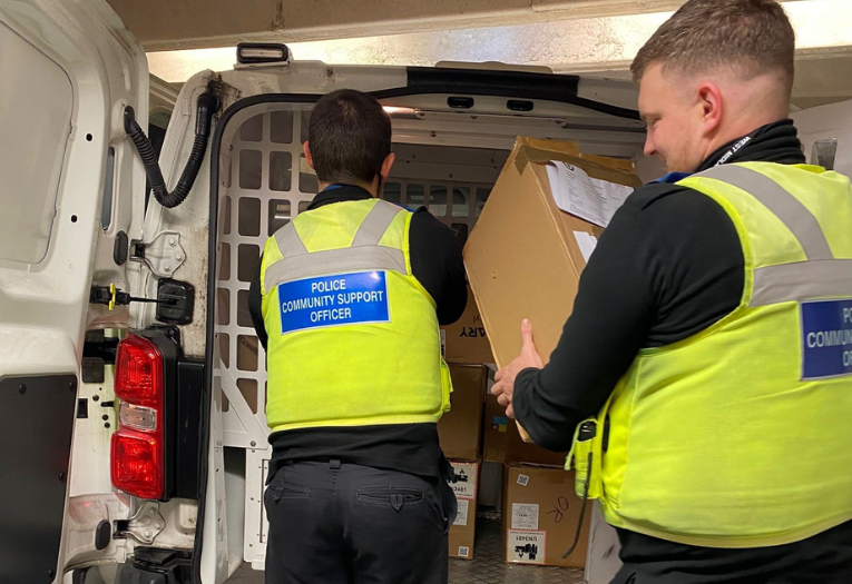 Image depicts two police community support officers placing seized boxes in the back of a police van.