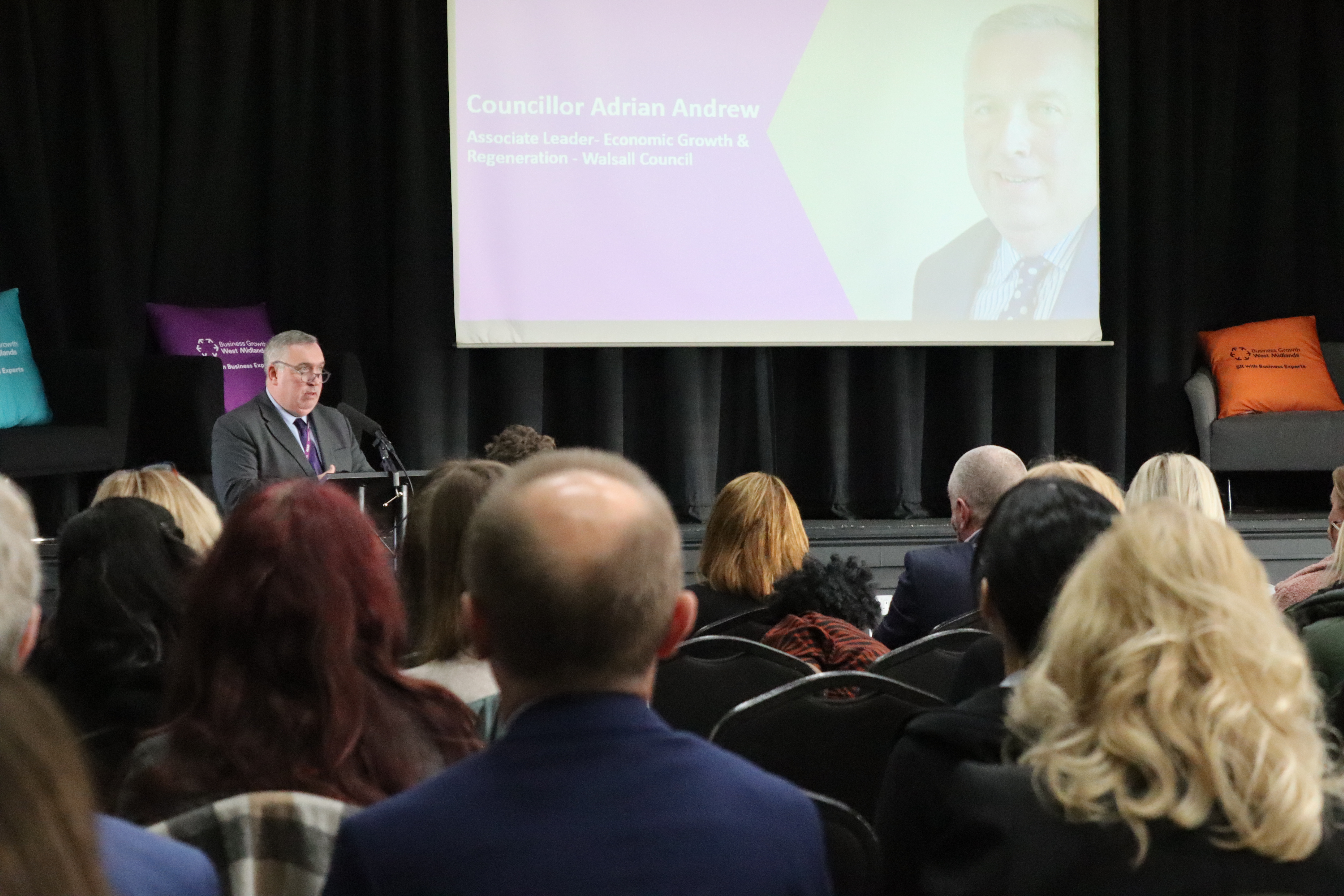 A crowd of people at an event looking towards a man in a suit who is speaking to the audience