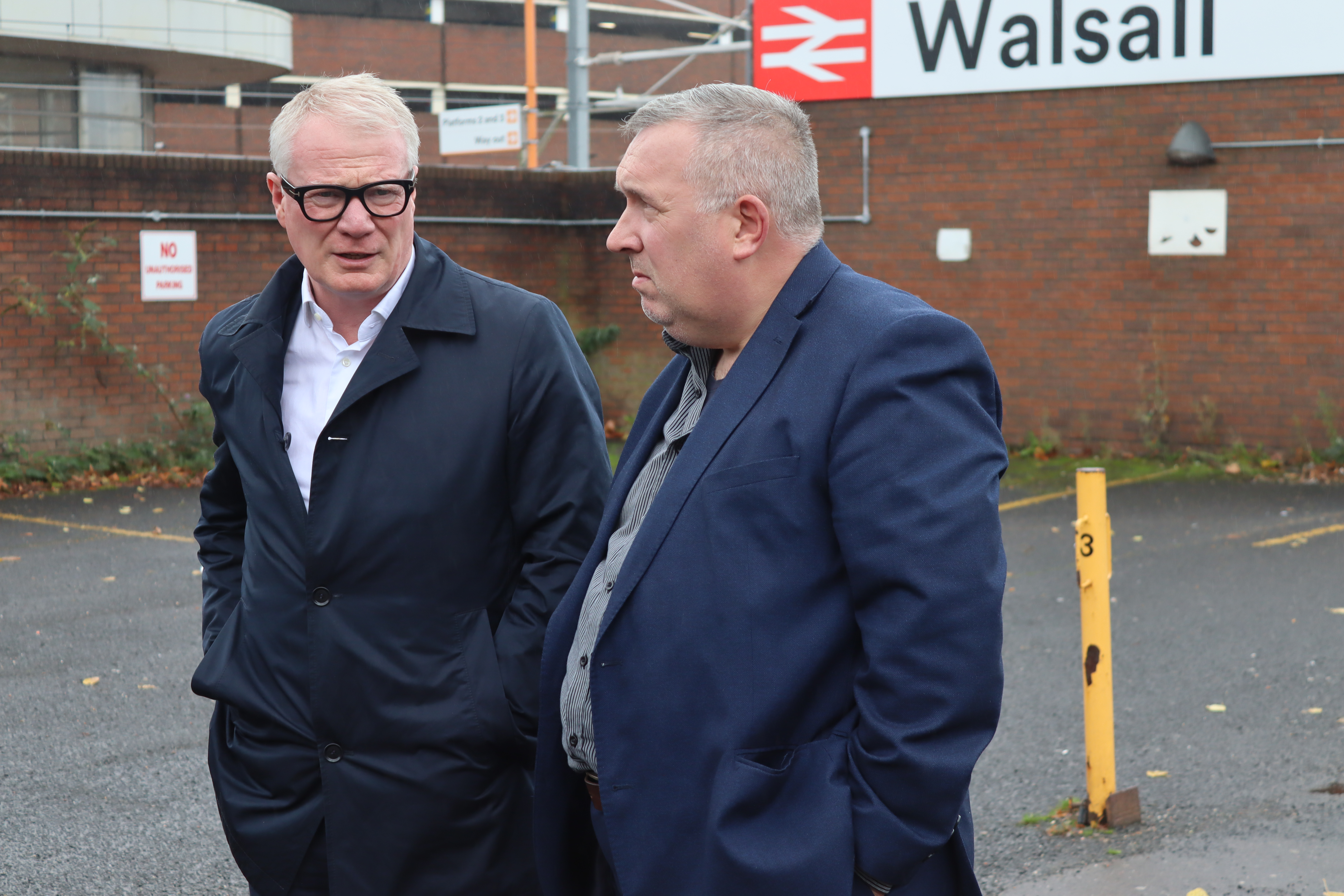 Mayor of the West Midlands Richard Parker and Councillor Adrian Andrew stand together outside Walsall train station, they are talking and both are wearing navy suits.