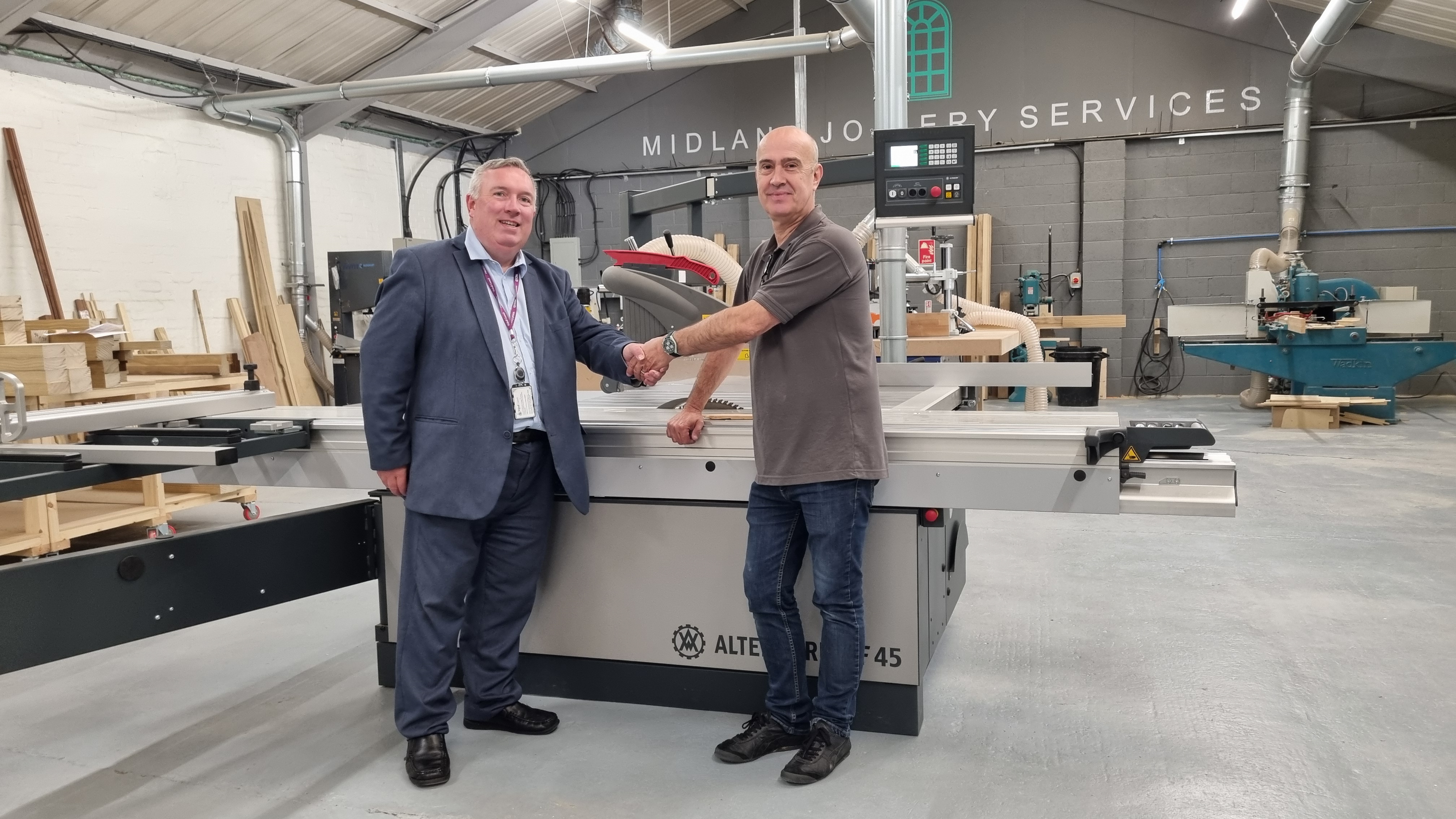 Two men shake hands, in front of a large piece of machinery, and smile at the camera