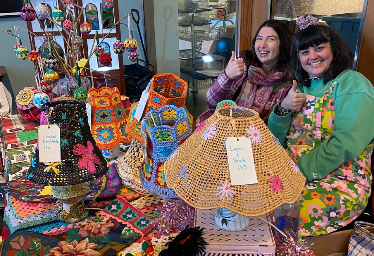 Two people smiling at a stall, surrounded by crocheted goods