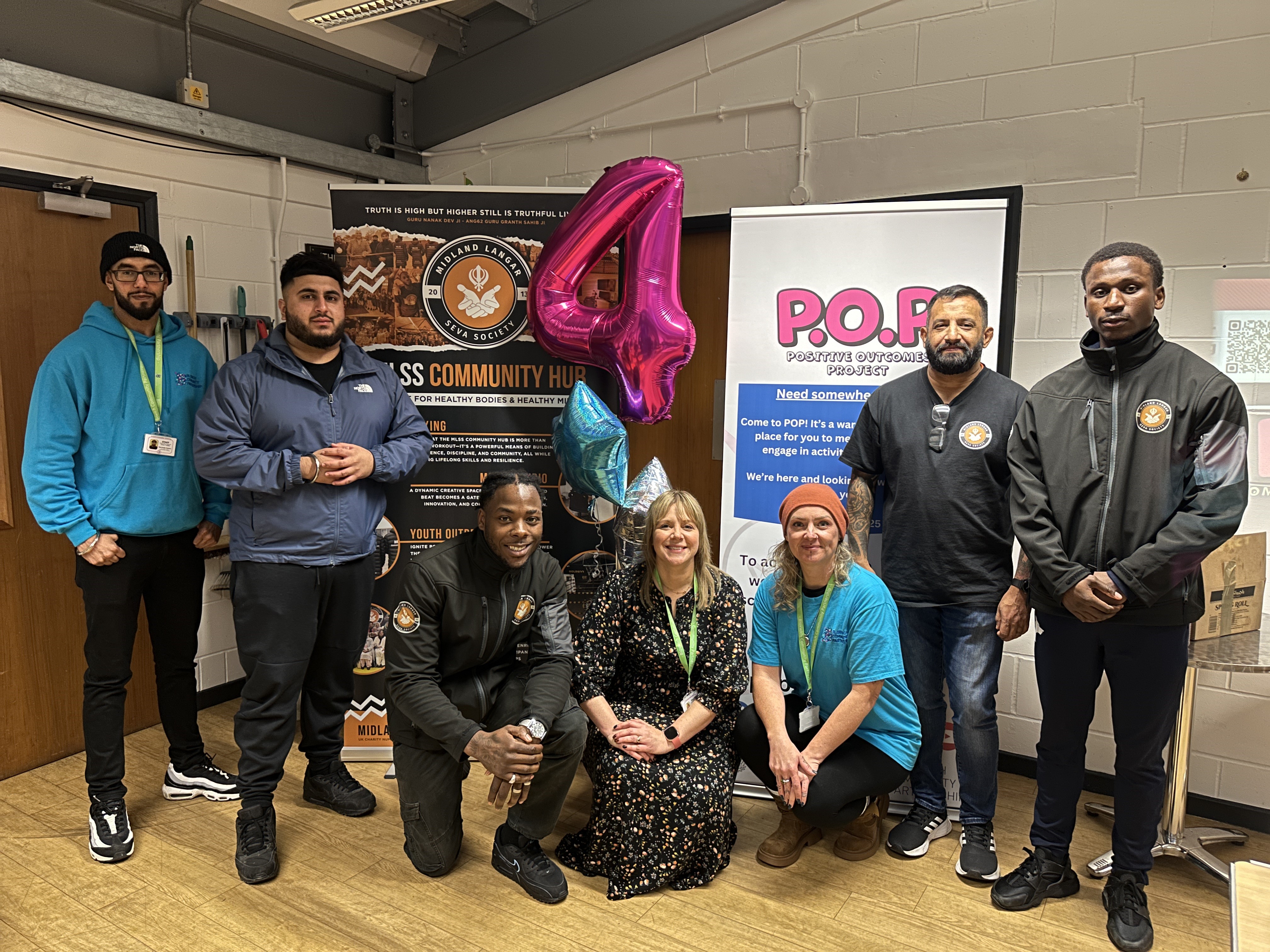 Image depicts seven representatives from Bloxwich Community Partnership and Midland Langar Seva Society in front of two banners and a number 4 balloon.