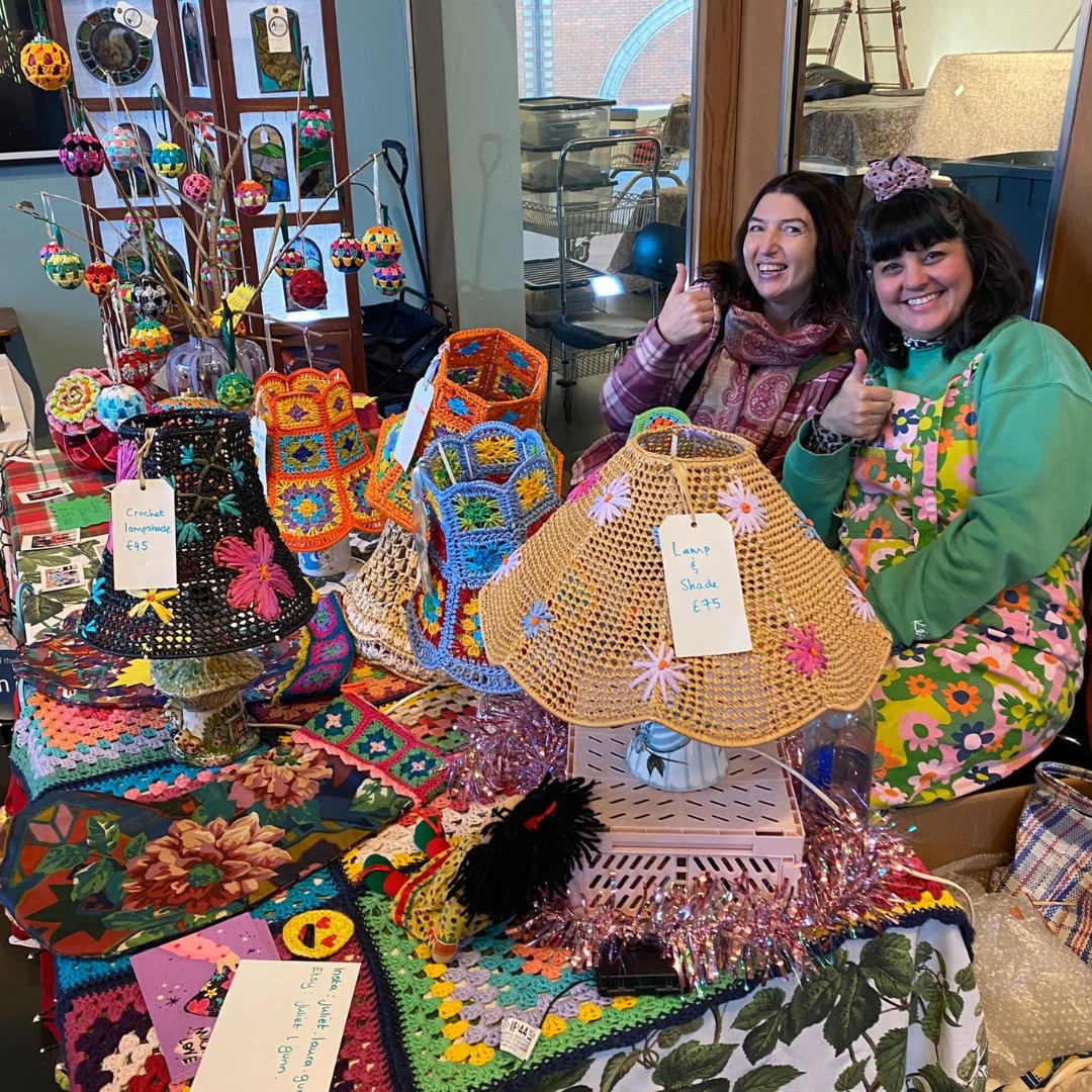 Two stallholders at a craft fair, with hand crafted items laid out on the table in front of them.