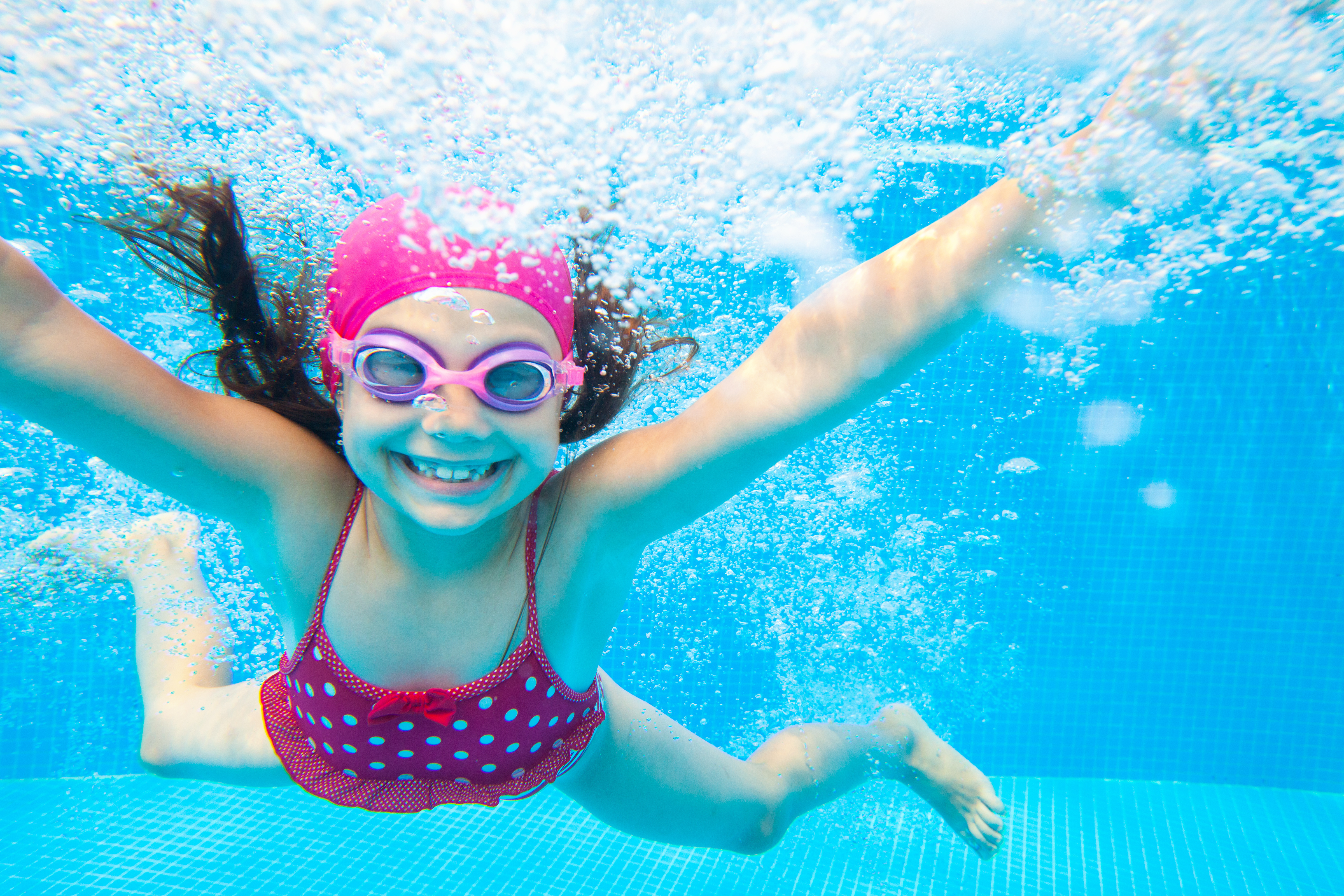 child with pink costume on and goggles underwater smiling 