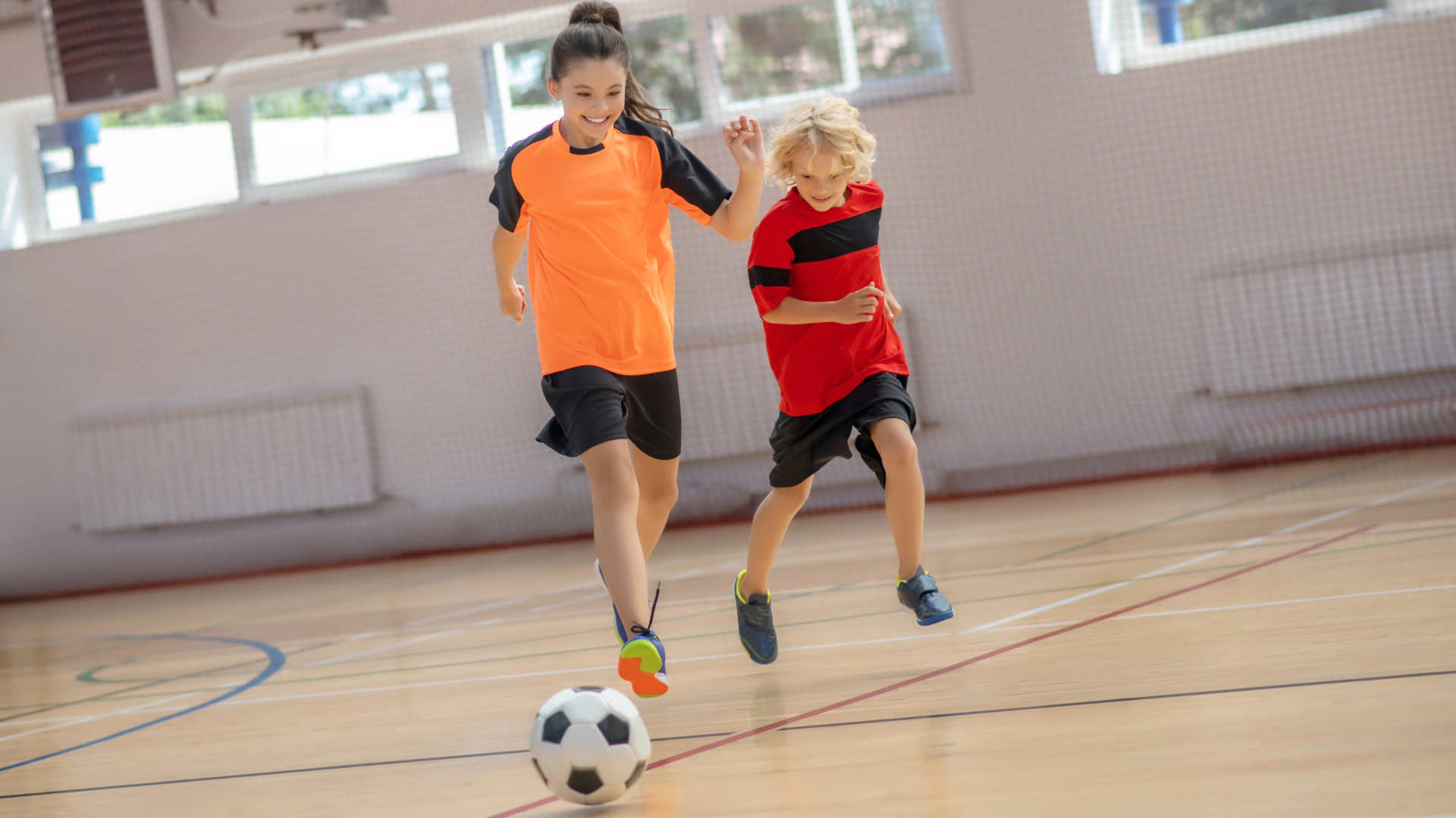 boy and girl kicking a football in an indoor sports hall smiling