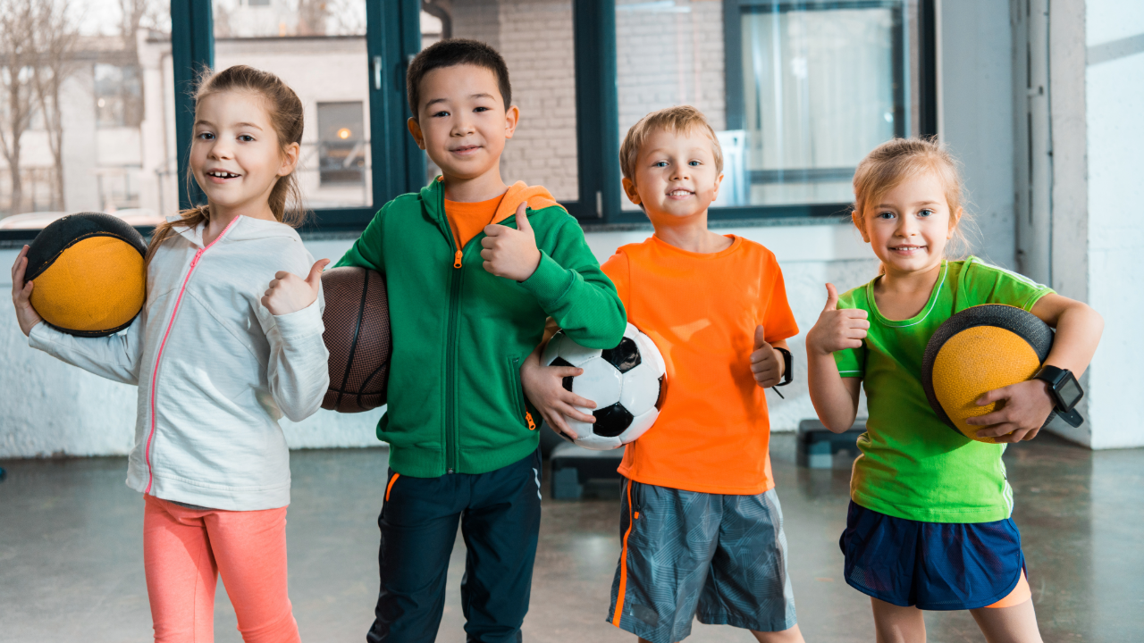 young children holding football, basketballs in an indoor sports hall with windows in the background 