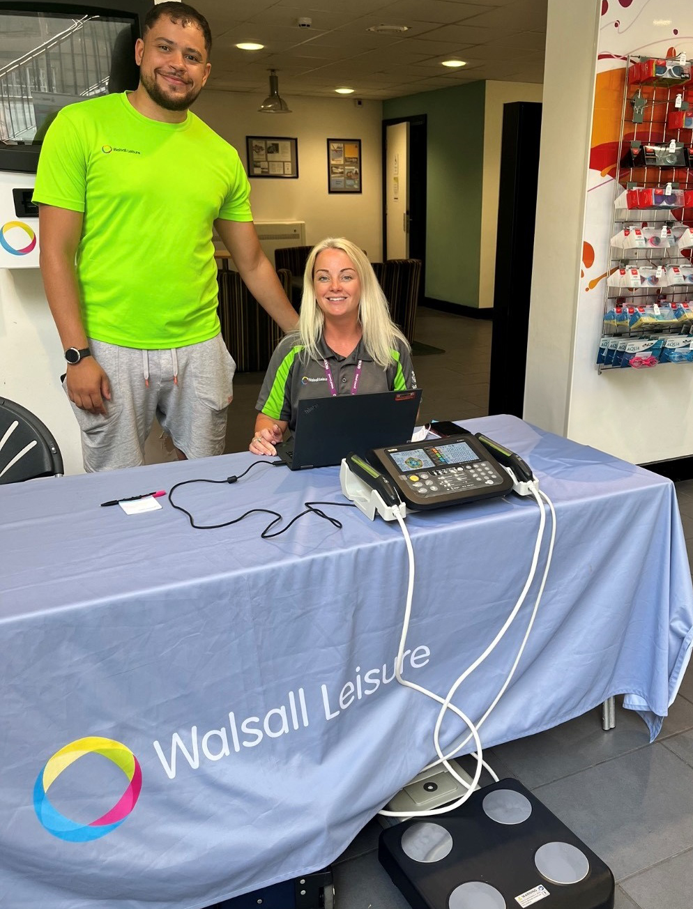 Female and male instructor sitting behind a desk with some weighing scales smiling in a leisure centre reception area