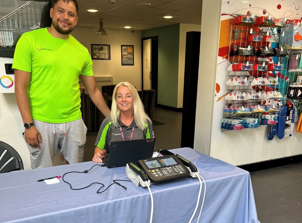 female and male instructor smiling behind a desk with a set of scales indoor in a leisure centre reception