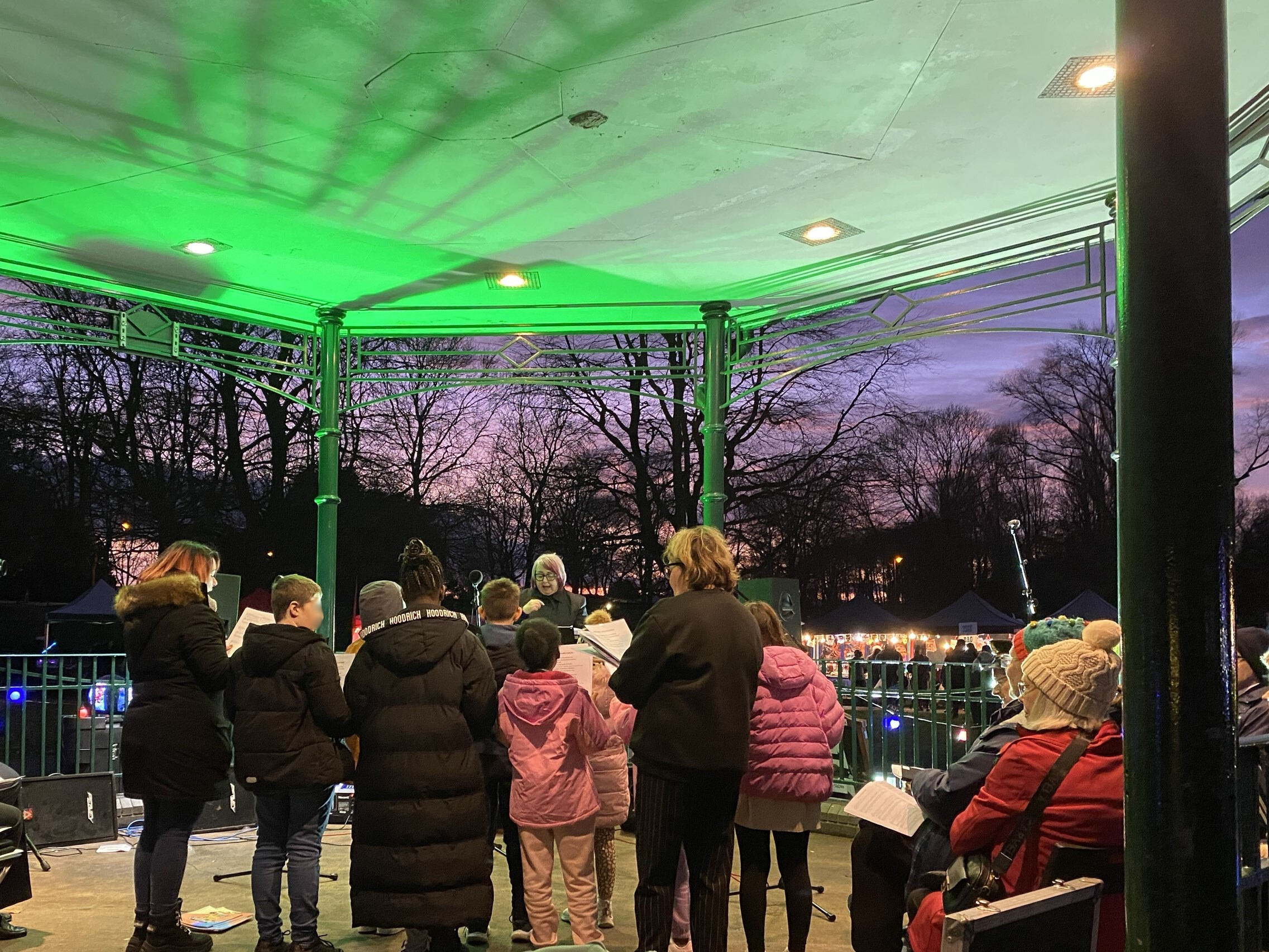 Image depicts a group of school children from behind singing at the bandstand at Willenhall Memorial Park.