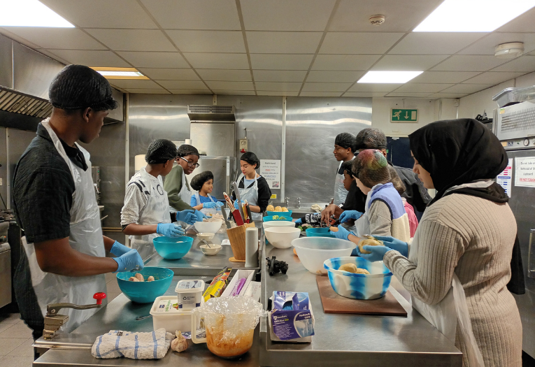 Image depicts a group of children and young people preparing food in a professional kitchen.