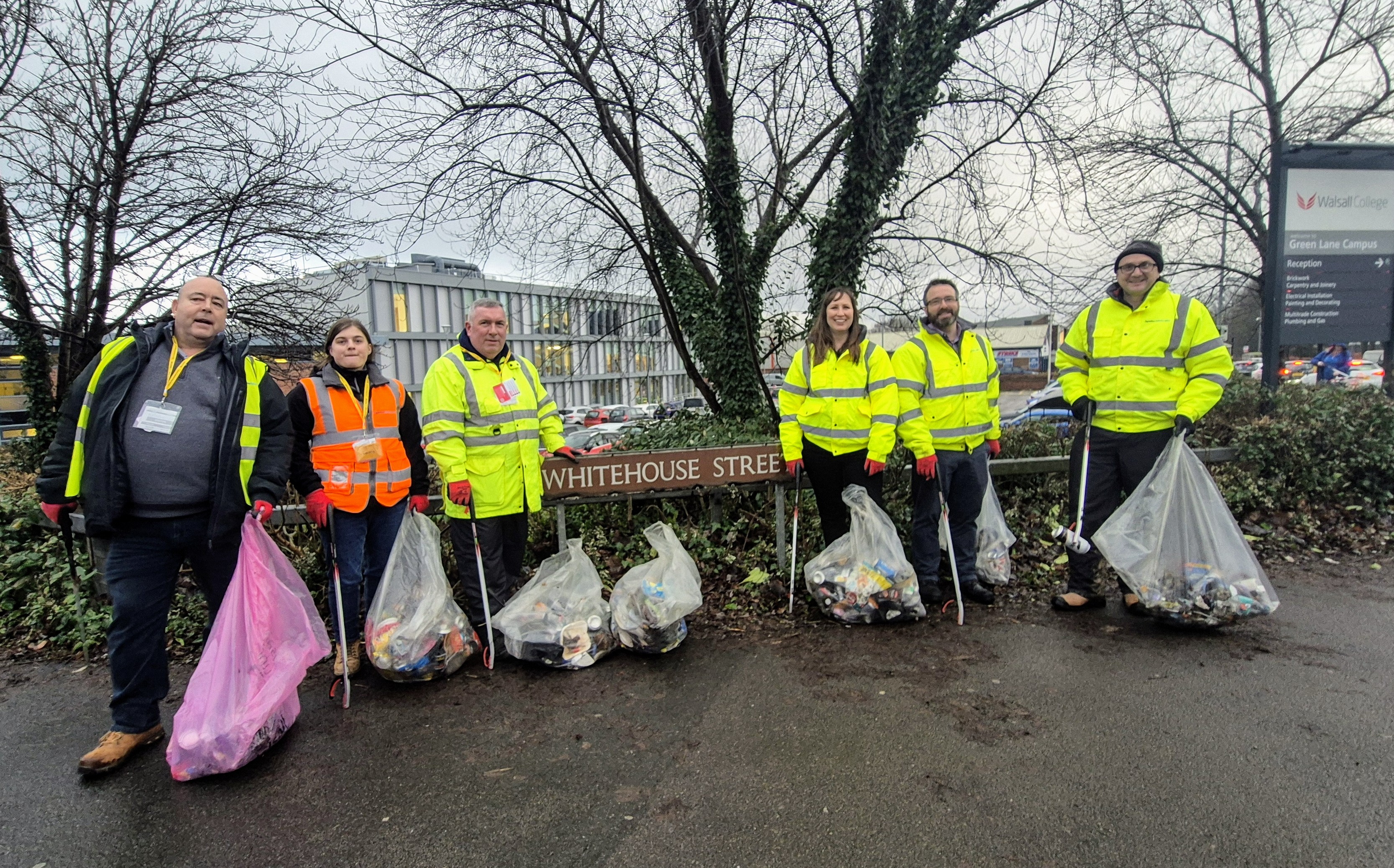 a group of 6 people in high vis jackets holding bags of litter stand around a street sign that says Whitehouse Street