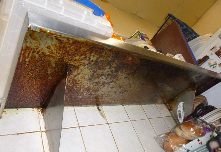 Image shows dirty / greasy underside to metal shelf in kitchen located to left hand side of fridge freezer.