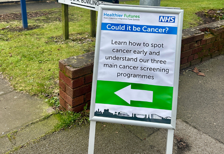 Image depicts a sign outside a community centre that reads 'Could it be Cancer? Learn how to spot cancer early and understand our three main cancer screening programmes'