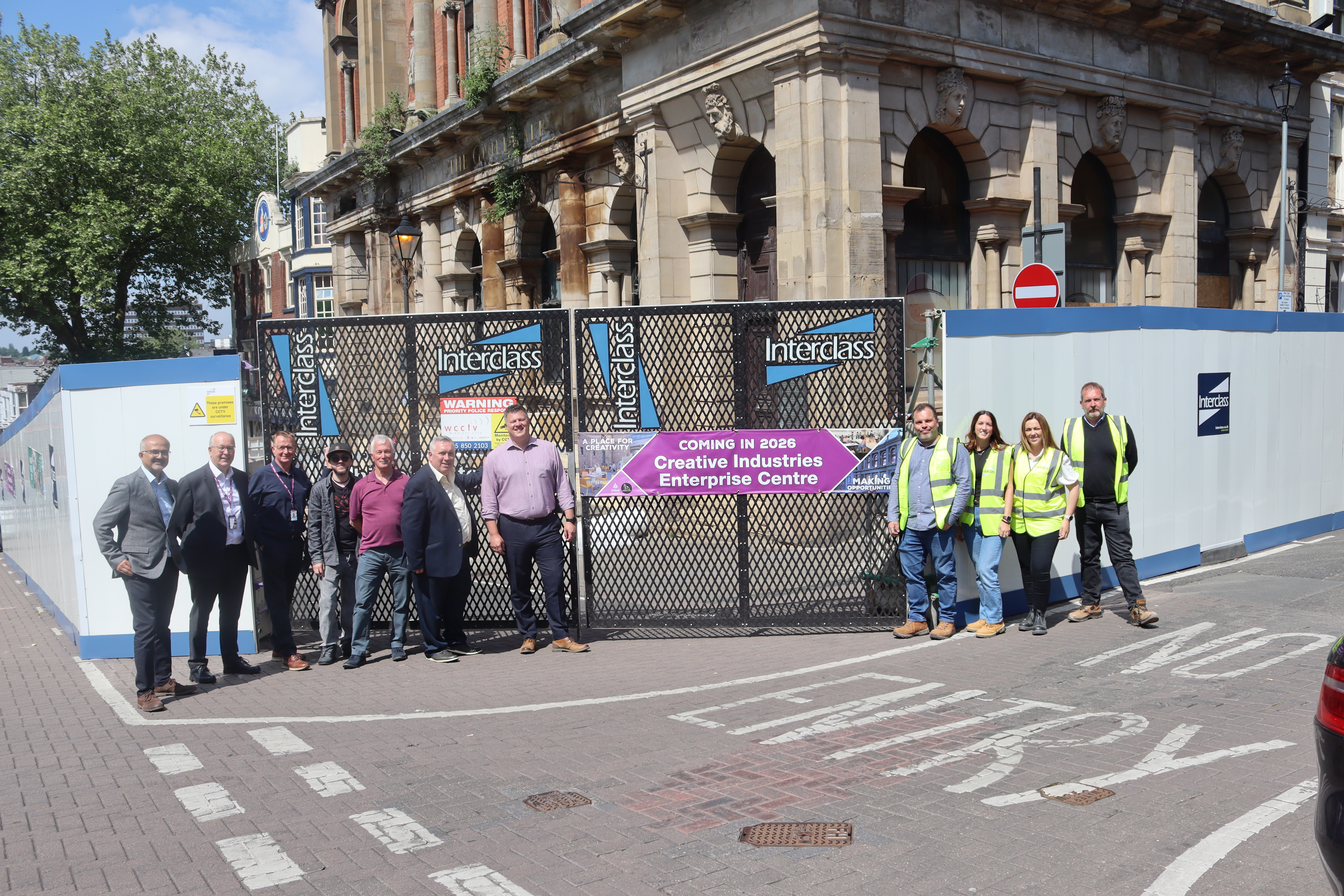 A group of people including contractors in high vis jackets stand in front of construction hoardings around the guildhall