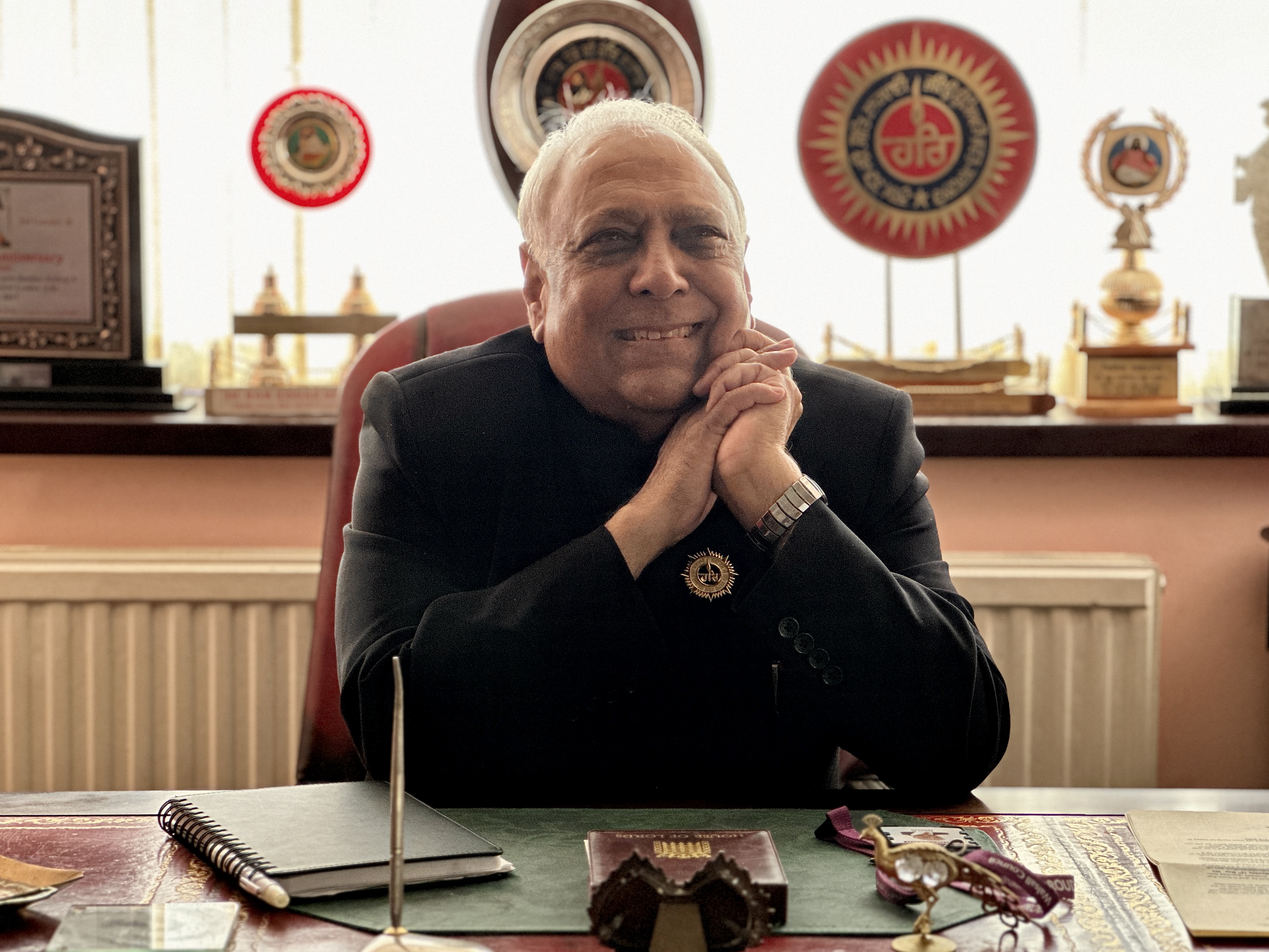 Image depicts Councillor Ram. K. Mehmi MBE smiling at his desk with his hands together on the side of his face.