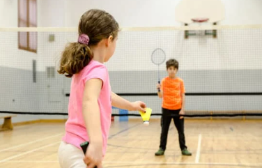 young girl in a pink top and blurred out boy in an orange top playing badminton in a sport shall 