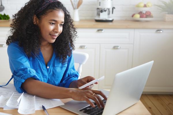 Woman sitting at a table in her kitchen, using a laptop to pay bills