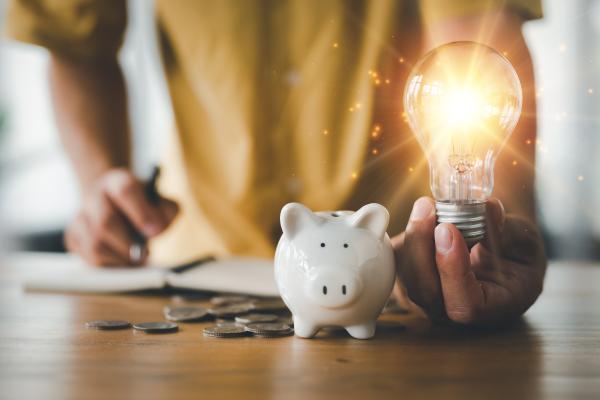 A person's hand holding a lit light bulb next to a piggy bank and coins