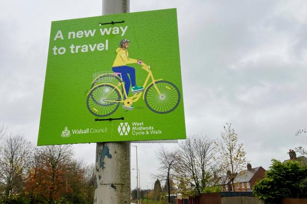 Image shows a green sign saying 'a new way to travel' on a lamp post.