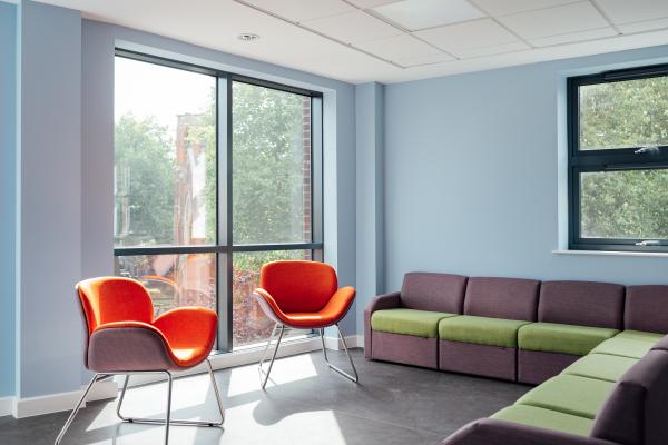 Inside a bright room with pale blue walls and large windows with green trees outside. Two bright orange chairs are infront of the window and there is a large grey and green corner sofa opposite