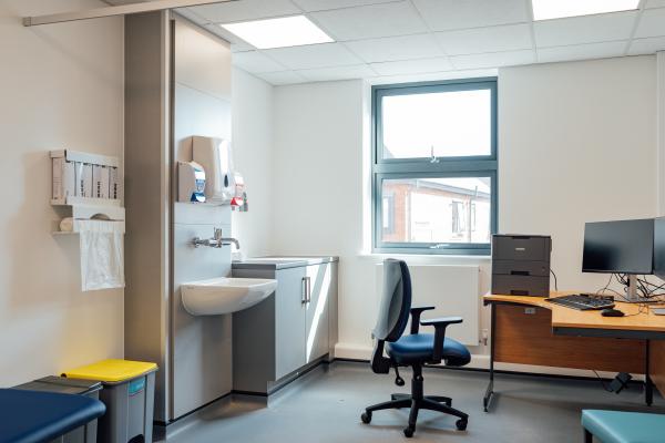 Inside a doctors office, there is a corner desk with a computer and blue office chair in the right hand corner next to a window. On the other side of the room is a sink and shelf of medical supplies