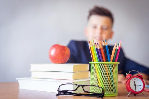 Image depicts glasses, pencil holder, red alarm clock, three books, an apple on a table with a child in school uniform in the background.