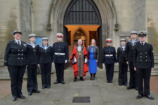 The mayor of walsall in red mayoral robes, next to the mayoress in a royal blue dress, stand smiling in the middle of a group of marine cadets in black uniforms outside a church entrance.