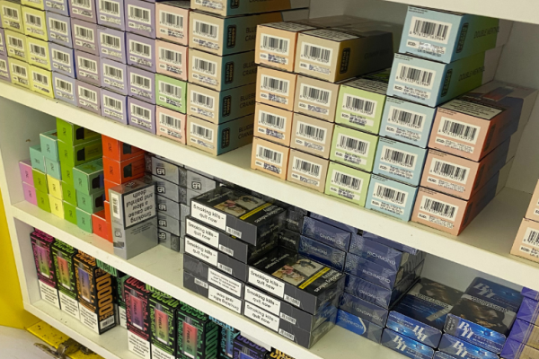 Image depicts illicit cigarettes and vapes concealed underneath a counter across three shelves.
