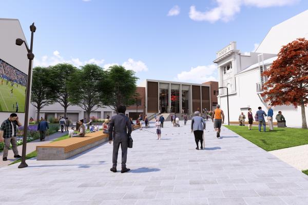 A new town centre square with a direct pedestrian route leading towards the large glass frontage of Rail station / Saddlers Centre entrance. There is grass either side of the walkway and a large screen on the side of a building on the left.