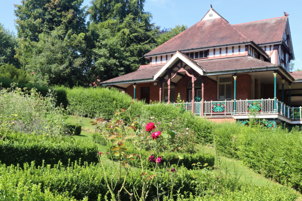 A Victorian building surrounded by a verandah with decorative railings, set in Walsall Arbortum showing the rose garden in front and trees behind