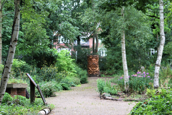 A view through the sensory garden, a path surrounded by trees, shrubs and plants leading to a wooden sculpture of a green man