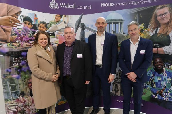 Four people dressed smartly smile whilst looking towards the camera, behind them is a large banner depicting a collage of Walsall landmarks with a purple strip above that reads Walsall Council