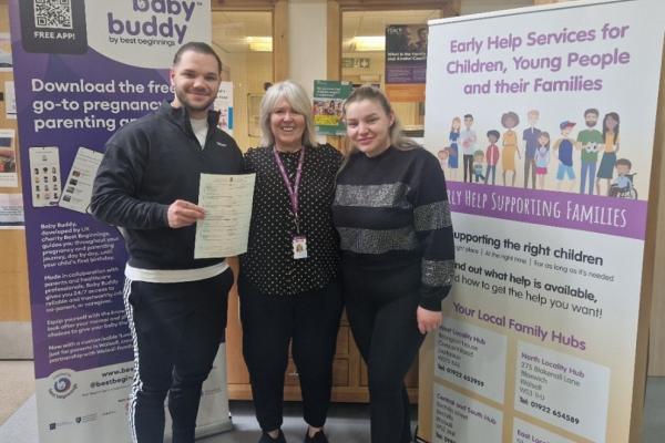 Baby Rares' mum and dad with Holly Holdsworth, Principal Registrar at Walsall Council. All three people are stood in front of pull up banners at the West Locality Family Hub.