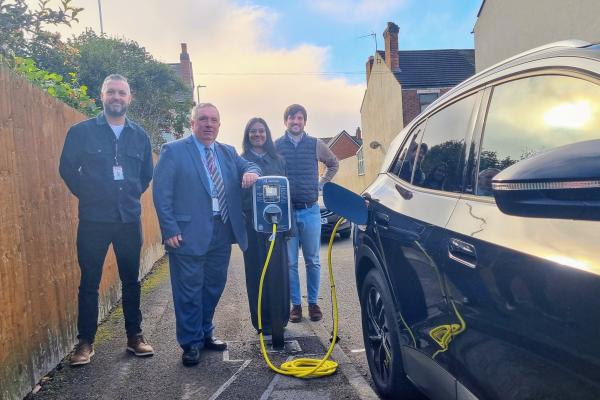 Four people stand next to a black car plugged into an electric vehicle chargepoint 